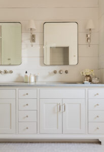 Elegant bathroom vanity with dual sinks, white cabinetry, and two mirrors flanked by wall-mounted lamps. Fresh flowers and bath products are on the countertop in this custom home.